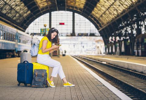 photo of a waiting woman in a train station seated on her luggage and using her mobile phone